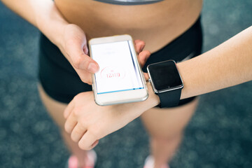 Young fitness girl checking her performance on smatphone and smartwatch after hard workout in gym.