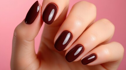 A hand with meticulously manicured dark brown nails is elegantly posed against a gentle pink backdrop, highlighting the glossy finish