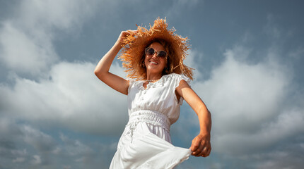 A Joyful Woman Wearing a Beautiful Summer Dress Stands Gracefully Against a Cloudy Sky Backdrop