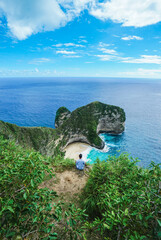 Man sitting close to the edge and looking to the incredible green rocks with beautiful tropical beach and deep blue sea on Kelingking Beach in Nusa Penida island, Bali, Indonesia. summer background  © Balnyes Visuals