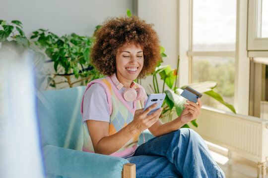 Happy curly hair woman doing online payment with credit card through smart phone at home
