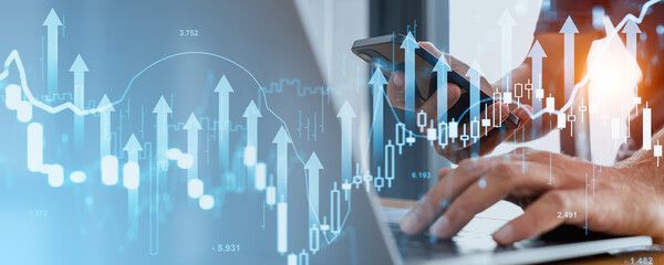 Hands holding a phone and typing on a laptop with financial charts and upward arrows overlaying the scene. Blue and white background. Concept of business trends and growth