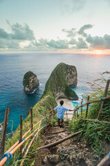 A young man is watching the sunset on a cliff overlooking the sea at Kelingking Beach, Nusa Penida, Bali. © Balnyes Visuals