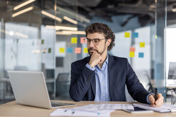 Businessman in suit working at desk with laptop and documents in modern office setting. Exhibits focus and deep thought. Concept of business, strategy, and technology.