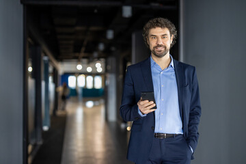 Confident businessman holding smartphone in modern office environment, displaying professional attitude. Smiling, walking in corridor, dressed in formal suit. Represents leadership, communication