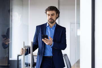 Serious businessman in blue suit uses smartphone while entering office meeting room. Focused professional engages in business communication, showcasing work environment and thoughtful expression.