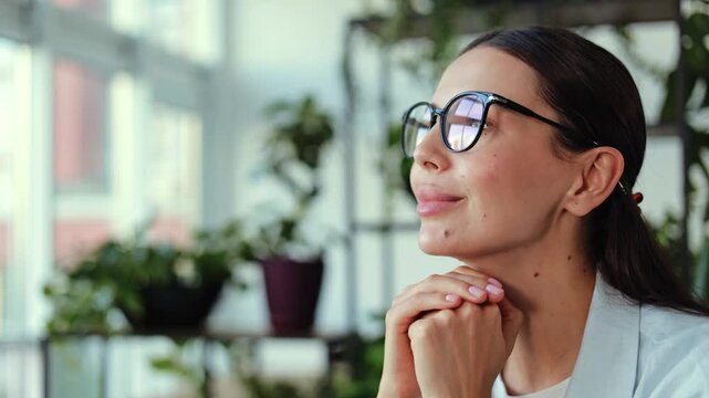 Close-up of young businesswoman with glasses in a green office, she is thoughtful and has her hands clasped under her chin. She is looking to the left of the frame and smiles.