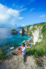 Young happy man traveller sitting on top of cliff and looking to Diamond Beach, Nusa Penida, Bali, Indonesia. summer background and summer vacation concept.