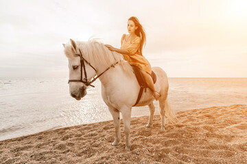 A woman in a dress stands next to a white horse on a beach, with the blue sky and sea in the...