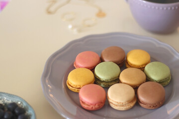 Plate of pastel macarons, cookies and chocolate, cup of tea of coffee, glass of bubble water, various berries, books and accessories on the table. Selective focus, pastel colors.