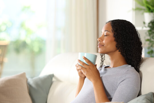 Black woman smelling hot drink on a sofa at home