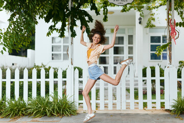 Happy young woman jumping in the air, wearing casual summer clothes, with a bright background featuring greenery and a white fence, conveying joy and positivity