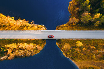 Aerial drone view of fall road and blue water lake sea ocean. Red car with a roof rack on country...