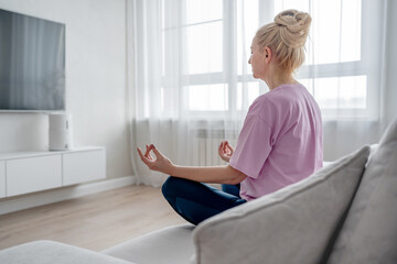 Elderly lady does yoga on the sofa in modern living room, enhancing mental and physical wellbeing....