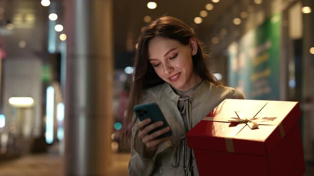 Excited Woman Holding Christmas Gift and Scrolling Apps on Smartphone Standing near Shopping Centre in the Evening