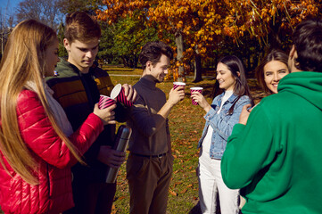 Smiling young people are walking in park. Group of best friends friends talking and drinking tea from paper cups on sunny autumn day. Autumn walk, friendship and leisure concept.