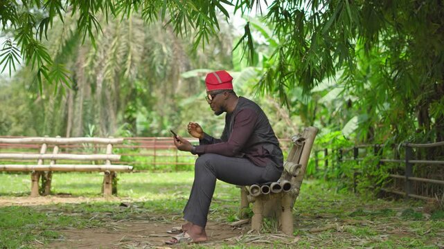 Full frame of a Nigerian Yoruba male in traditional attire and cultural cap video calls in a garden.
