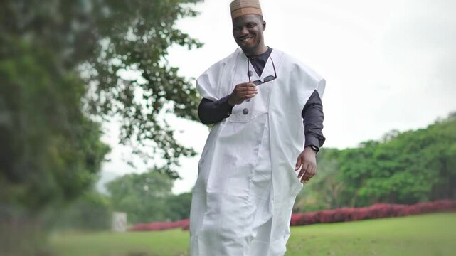 Slo-mo of a Nigeria Hausa wearing a white and black Babariga attire removes his sunglasses and smiles to the camera walking beautiful outdoor.