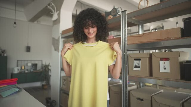 Cute Caucasian girl standing inside factory and placing on top of her clothes yellow t-shirt. Trying out new look. Thinking about changing stile while smiling to herself. Looking at camera.