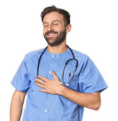 Hispanic male nurse in studio laughs out loudly keeping hand on chest.