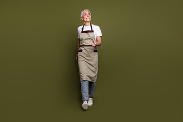 Full length photo of cheerful dreamy lady dressed apron owning cafe walking looking empty space isolated green color background