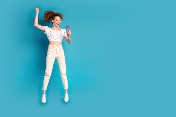 Full body photo of attractive young woman jump winning hold device dressed stylish white clothes isolated on blue color background