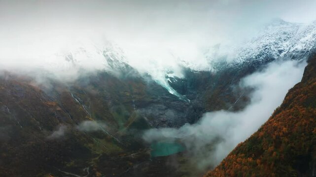 A fog-covered glacier descends through the mountain, with water trickling from its edges amid fall colors. Aerial view.