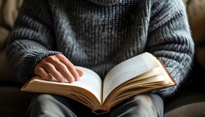 Cozy reading session on a couch with hands gently resting on book pages in a warm gray sweater