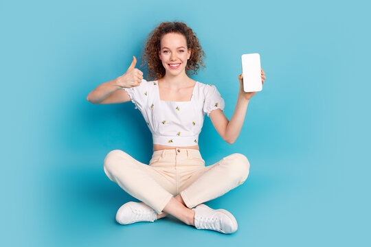 Full length photo of lovely young lady sit floor gadget qr code thumb up dressed stylish white garment isolated on blue color background