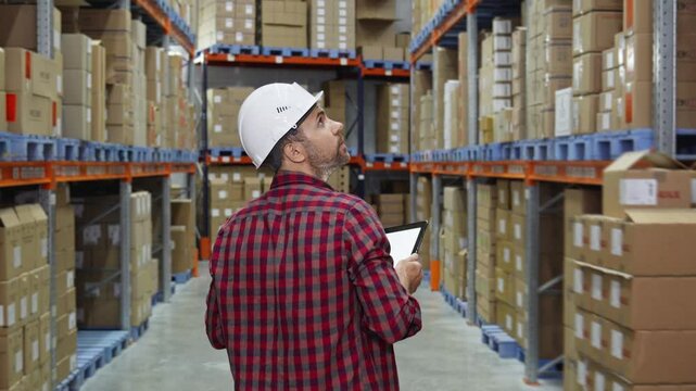 Worker in hard hat surveys rows of neatly stacked boxes in warehouse. Rear view of male worker in white hardhat inspecting goods in warehouse. Tracking shot in 4K, UHD