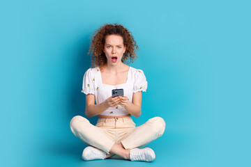 Full length photo of lovely young lady sit floor gadget frightened dressed stylish white garment isolated on blue color background