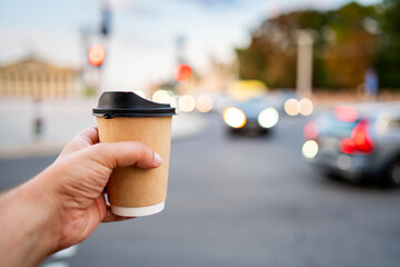 A close-up of a hand holding a takeaway coffee cup with a black lid, against a blurred city street background. The cup is white with a simple design, perfect for illustrating urban lifestyle