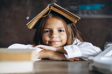 Little schoolgirl sitting at the desk in classroom having a book on her head