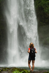 Obraz premium A young blonde girl, a traveler, admires a majestic waterfall with a backpack.