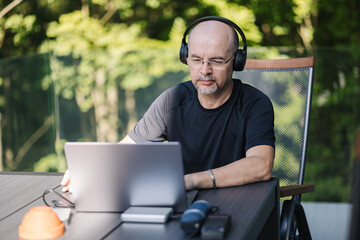 A mature man with works remotely from home while wearing headphones and glasses. Man 