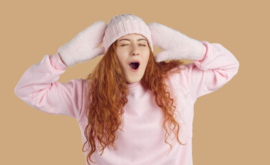 Funny young woman with long curly wavy ginger hair in cozy warm light pink hat, fluffy mittens and comfortable sweatshirt stretching her arms and yawning on beige background. Winter fashion concept