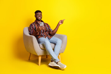 Full body portrait of nice young man sit chair phone direct finger empty space wear shirt isolated on yellow color background