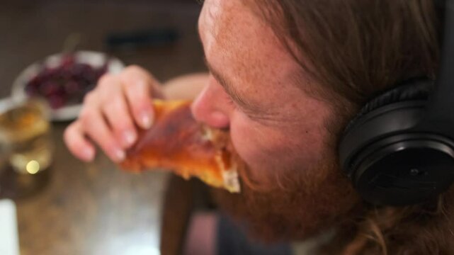 Top view of gluttonous redhead millennial man eating delicious fresh bun and listening to music close up.