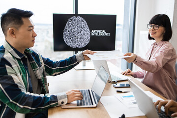 Team members collaborating on artificial intelligence project in modern office environment. Two individuals sharing documents and ideas while working on laptops, focusing on innovative technology.