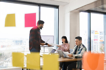 Team of creative architects collaborating in modern office with bright sticky notes on window. Casual business meeting exchanging ideas.