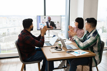 Team video conference meeting in modern office with participants applauding virtual presenter. Colleagues gathered around table with laptops, taking notes, and engaging in discussion.