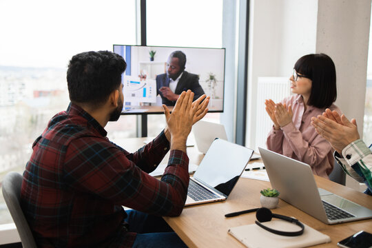 Business team clapping during video call with remote colleague. Office workers engaged in virtual conference, applauding performance and collaboration. Modern workspace and technology