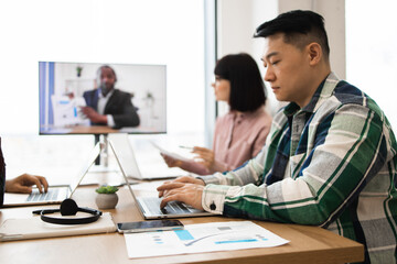 Professionals collaborating during team meeting with virtual presentation. Participants focused on laptops while engaging with remote presenter. Concept of hybrid work, teamwork, and technology.