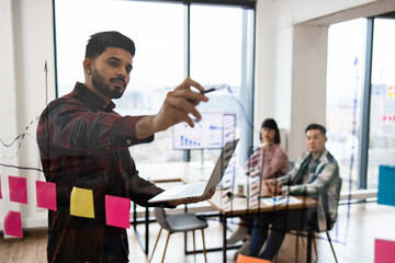 Office meeting featuring brainstorming session with colleagues. Man writing on glass wall with sticky notes and graphs in background. Team collaboration and business strategy planning in workspace.