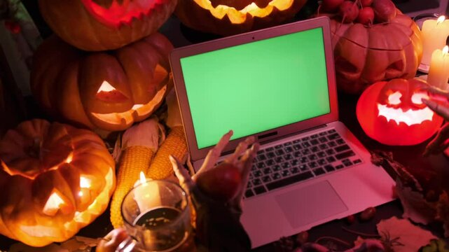 Creepy monster hands next to laptop with green screen surrounded by shimmering pumpkins jack o lanterns, halloween holiday background.