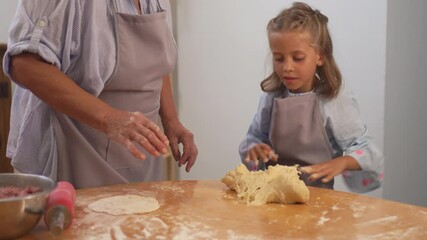 Cute girl kneading pastry dough in assistance of unrecognizable grandmother on table at home. Little female child is learning baking from elderly woman, baking concept 