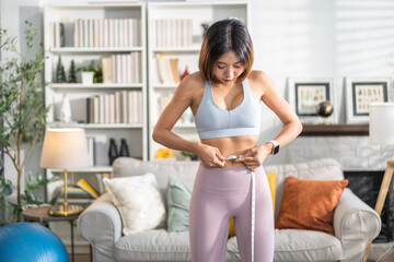 Asian woman measuring waist with tape while smiling in sportswear, fitness and weight loss concept,...