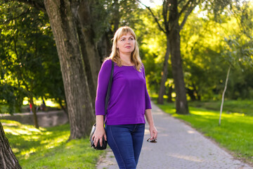 A woman in her 40s, wearing a purple blouse, walking through a park surrounded by nature. Blurred