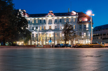 Naklejka premium Beautifully illuminated historic building in the heart of downtown with motion blurred people at night. Chill destinations travel trend. Vilnius, Lithuania. Copy space