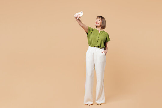Full body elderly blonde woman 50s years old wear green shirt casual clothes doing selfie shot on mobile cell phone post photo on social network isolated on plain beige background. Lifestyle concept.